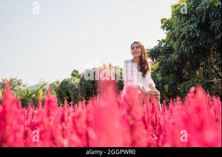 Allegro bella giovane donna asiatica seduta con braccia di sollevamento in fiore cockscomb in giardino Foto Stock