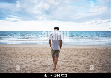 Posteriore di uomo solitudine in camicia bianca che cammina sulla spiaggia in mare Foto Stock