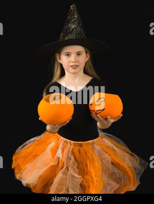 Una ragazza allegra con i capelli lunghi e in un costume di strega per la celebrazione di Halloween tiene fuori due zucche Jack. Cappello nero con ciottoli e blusa, arancione Foto Stock