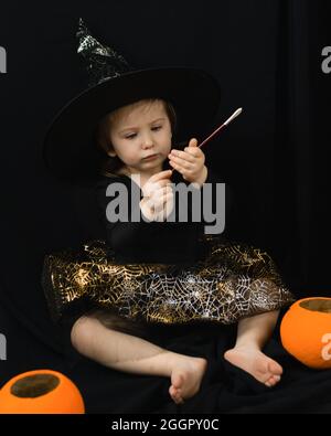 Una bambina in un costume da strega - un cappello e una gonna con una rete di ragno - si siede su uno sfondo nero ed esamina intentemente una bacchetta magica. Accanto è un Foto Stock