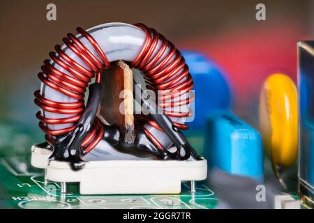 Induttore con nucleo in ferrite su supporto bianco e condensatori colorati su scheda a circuito stampato verde. Primo piano della bobina toroidale elettronica con filo di rame isolato. Foto Stock