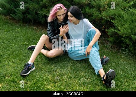 due ragazze adolescenti guardano qualcosa su uno smartphone, sull'erba dietro i cespugli del parco Foto Stock