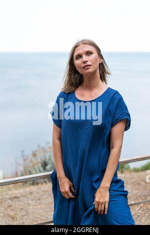 Donna carina si erge in un abito blu sullo sfondo del mare e del cielo. Espressione facciale pensiva. Gode della natura e dell'aria pulita Foto Stock