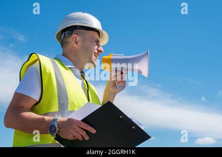 Il costruttore in un casco bianco urla in un megafono contro uno sfondo blu del cielo Foto Stock