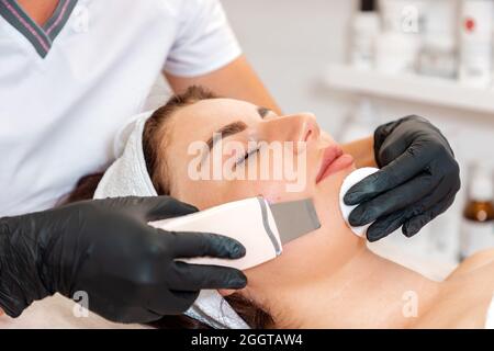 L'estetista in guanti di gomma pulisce il trucco del cliente utilizzando un dispositivo a ultrasuoni. Primo piano della faccia. Concetto di procedure di bellezza e cosme professionale Foto Stock