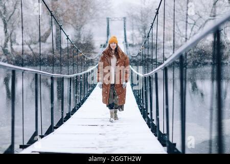 Una donna cammina sul fiume su un ponte sospeso in inverno. Giovane ragazza in abiti caldi si trova su un ponte di legno in una fredda giornata innevata. Foto Stock