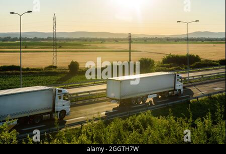 White Trucks con semirimorchio che guida lungo l'autostrada sul cielo al tramonto e sullo sfondo rurale. Servizi e logistica dei trasporti Foto Stock