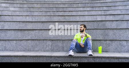 L'elegante uomo hipter con barba si siede da solo sui gradini. Ragazzo in abiti casual alla moda e luminosi. Banner Foto Stock