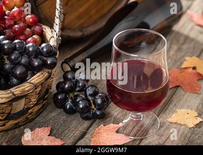 Bicchiere di vino rosso con uva, bottiglia di vino e barile su tavola di legno esterna con foglie autunnali Foto Stock