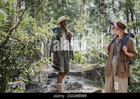 Coppia anziana interrazziale con zaini sorridenti mentre camminano nella foresta Foto Stock