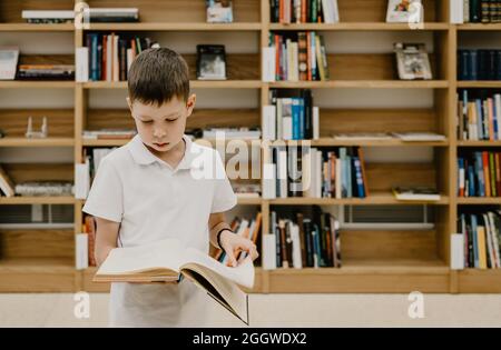 Un ragazzo si alza in biblioteca e legge un libro mentre è in piedi. Preparazione per i compiti. Il ragazzo ama leggere. Spazio libero a scuola. Extracurricolare Foto Stock