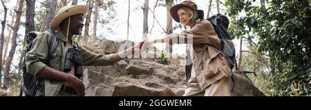 Sorridente uomo afroamericano che tiene la mano di moglie con zaino in foresta, banner Foto Stock