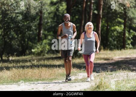 Sportivo afroamericano con bottiglia di sport che corre vicino felice moglie in foresta Foto Stock