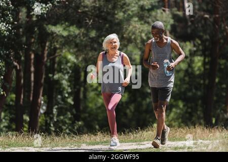 Coppia multiculturale allegra con bottiglia sportiva che corre nella foresta Foto Stock