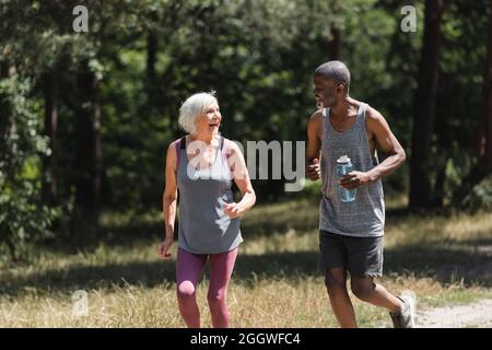 Allegra coppia multietnica con bottiglia d'acqua che corre nella foresta Foto Stock