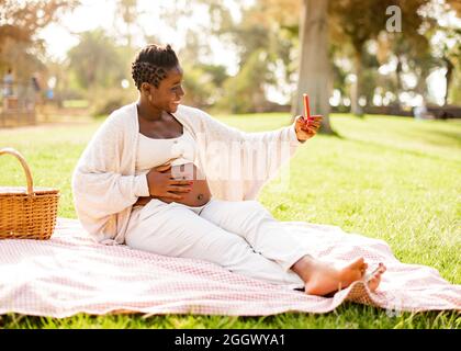 Donna afroamericana incinta che prende selfie nel parco Foto Stock
