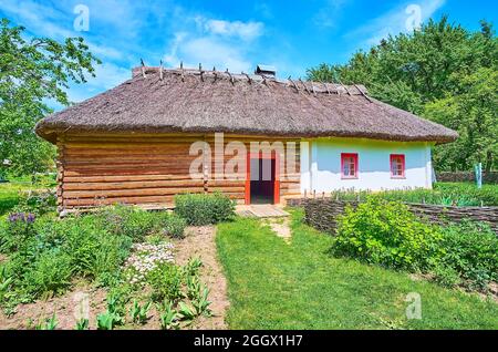 PEREIASLAV, UCRAINA - 22 MAGGIO 2021: Un giardino con fiori in fiore e prato di fronte alla casa in legno hata con tetto di paglia e livina imbiancata Foto Stock