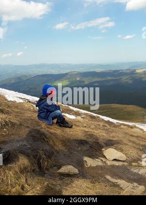 Il ragazzino si siede su una montagna sullo sfondo di altre montagne e del cielo blu Foto Stock