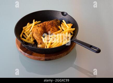 Vista ad angolo alto della gamba di pollo arrosto con patatine fritte in padella in ghisa su piastra di legno. Riflessione su tavolo di vetro Foto Stock