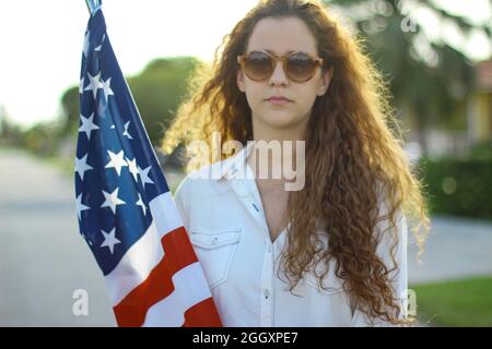 Giovane donna caucasica e ispanica rossa che indossa occhiali da sole tenendo la bandiera americana fuori. Foto Stock