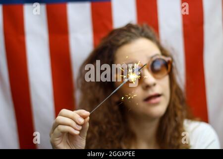 Giovane rosso ispanico e caucasico che tiene uno scintillio in piedi di fronte alla bandiera americana. Foto Stock