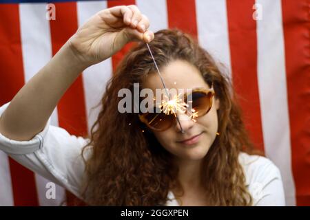 Giovane rosso ispanico e caucasico che tiene uno scintillio in piedi di fronte alla bandiera americana. Foto Stock