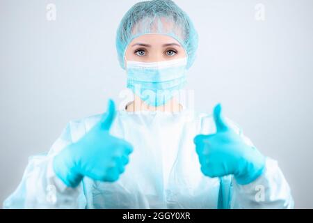 un'infermiera in un abito medico, maschera e guanti protettivi dà un pollice su Foto Stock
