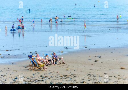 Un gruppo di amici si siede in sedie a sdraio sulla spiaggia di Saundersfoot a Pembrokeshire, galles, mentre le famiglie pagaiano in mare alle spalle. Foto Stock