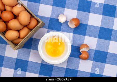 Una piccola cassa di uova fresche con due uova screpolate poggia su un tavolo coperto in una tovaglia tradizionale a scacchi blu e bianco. Foto Stock