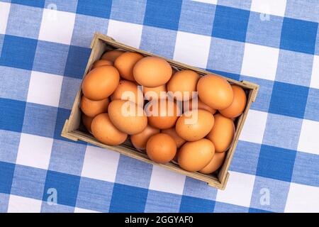 Una piccola cassa di uova di gallina marroni raccolte di fresco si riposa su una tradizionale tovaglia da picnic a scacchi blu e bianco. Foto Stock