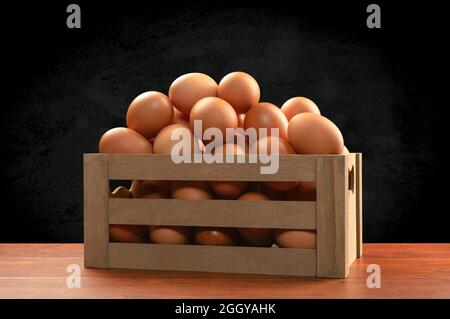 Una piccola cassa marrone di legno di uova appena raccolte sulla parte superiore di un tavolo di teak e incorniciata da un sottile sfondo nero, chiazzato. Foto Stock