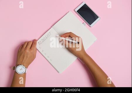 Disposizione piatta delle mani femminili che tengono la matita, scrivendo nel taccuino, controllando l'elenco delle cose da fare. Telefono cellulare su sfondo rosa con spazio per il testo. Affari Foto Stock