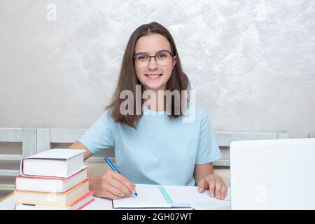 Uno studente adolescente sorridente seduto a un tavolo con libri sta scrivendo un'attività in un notebook. Concetto di ritorno a scuola. Concetto di scuola domestica. Foto Stock