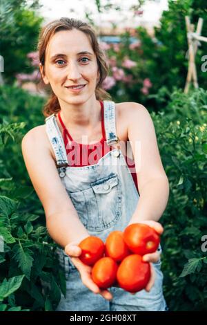 Ritratto di una donna attraente con pomodori rossi nelle sue mani. Le verdure appena tagliate delle piante piene di vita. La ragazza vegana prende il suo quotidiano f Foto Stock