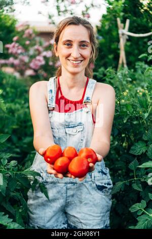 donna con verdure in mano guarda la macchina fotografica con un sorriso. Il coltivatore raccoglie il raccolto nel campo. Pomodori rossi da mangiare organicamente. Foto Stock