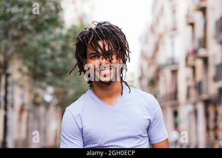 ritratto orizzontale di un giovane ispanico con i dreadlocks. Guarda la macchina fotografica e sorride mentre cammina per le strade di Barcellona Foto Stock