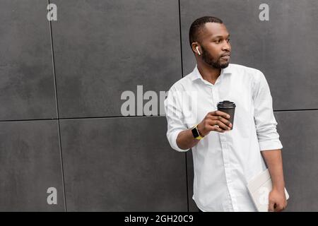 Elegante uomo d'affari afroamericano in piedi in camicia bianca, tenendo una tazza di caffè, avendo una pausa dopo conferenza di lavoro in linea. Bel maschio Foto Stock