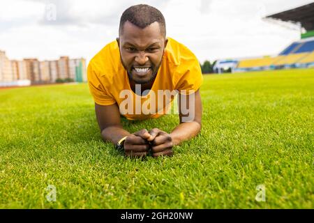 Ritratto di un maschio afro-americano stanco che lavora duro su un campo di erba allo stadio. Giovane atletico uomo che fa esercizio asse all'aperto in giallo Foto Stock