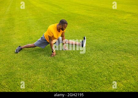 Uomo sportivo concentrato in un abbigliamento sportivo elegante che cerca di fare una spaccatura, seduto allo stadio allungando le gambe. Lavoro sportivo maschile afroamericano Foto Stock