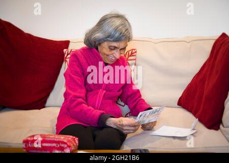 Donna indiana più anziana o anziana regalo di Natale di apertura o regalo e carta, seduto a casa. Donna anziana asiatica nel suo 80, Regno Unito Foto Stock