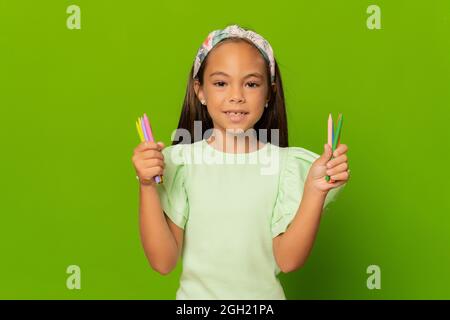 Ragazza bambino allegra che tiene matite di colore su sfondo verde. Istruzione e concetto scolastico. Foto Stock