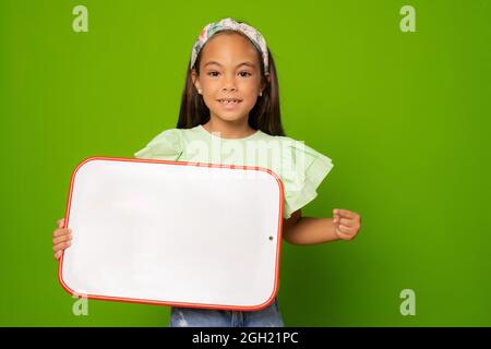 Ragazza bambino allegra che tiene la lavagna su sfondo verde. Istruzione e concetto scolastico. Foto Stock