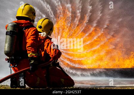 Squadra di due vigili del fuoco che indossa uniforme piena con serbatoio di ossigeno sul retro che tiene il tubo ha sparso l'acqua e faccia al fuoco caldo arancione grande con coraggioso e. Foto Stock