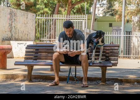 Tel Aviv, Israele - 16 agosto 2021: Un giovane seduto su una panca del parco con il suo cane, guardando il suo smartphone. Il cane sembra fare lo stesso. Foto Stock