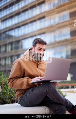 Foto verticale di un uomo che usa un computer portatile all'aperto. Uomo d'affari con un bel sorriso lavora a distanza per strada. Foto Stock
