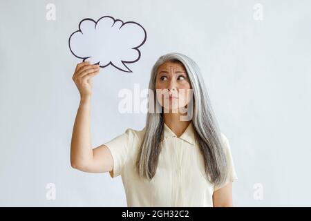 La donna matura in dubbio tiene la bolla di testo in bianco in piedi su sfondo chiaro Foto Stock