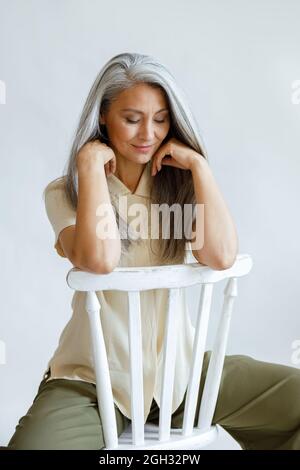 Donna asiatica di mezza età con capelli grigi si siede indietro sulla sedia su sfondo chiaro Foto Stock