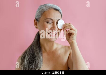 Positivo grigio capelli Asian donna rimuove il trucco da occhio con tampone di cotone in studio Foto Stock