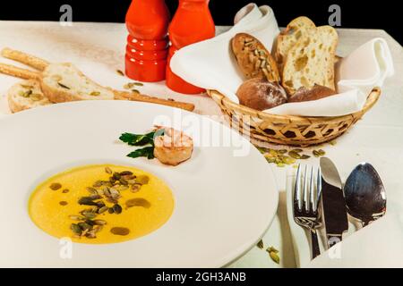 Zuppa di zucca cosparso di semi di zucca, gamberi in composizione con pane, posate, grissini su un tavolo Foto Stock