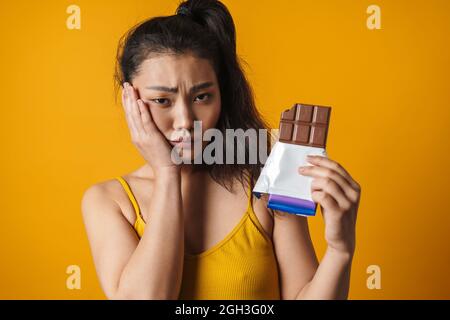 Ritratto di una giovane donna asiatica sconvolto che tiene la barra di cioccolato in piedi su sfondo giallo Foto Stock